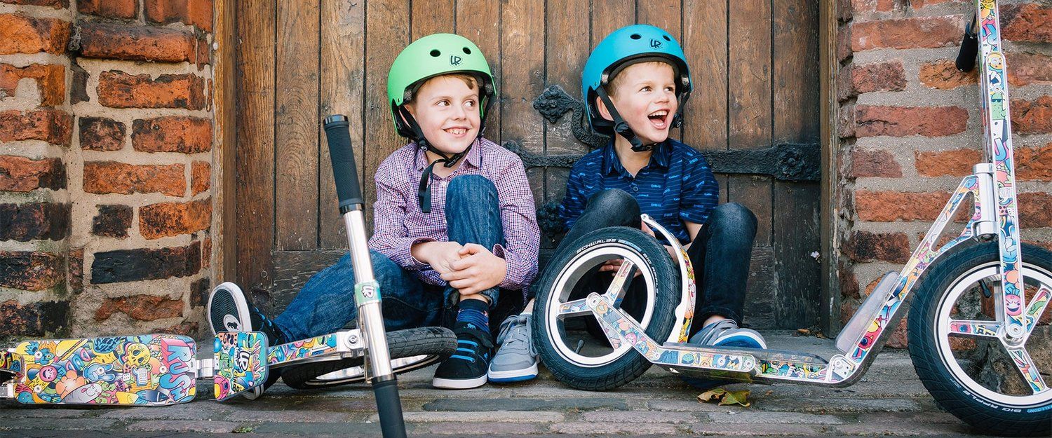 Two children sitting on the ground with scooters, wearing helmets, in front of a wooden door.