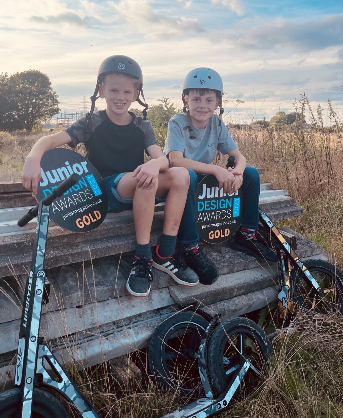 Two children with scooters, holding 'Junior Design Awards' gold award plaques, sitting on a wooden bench outdoors.